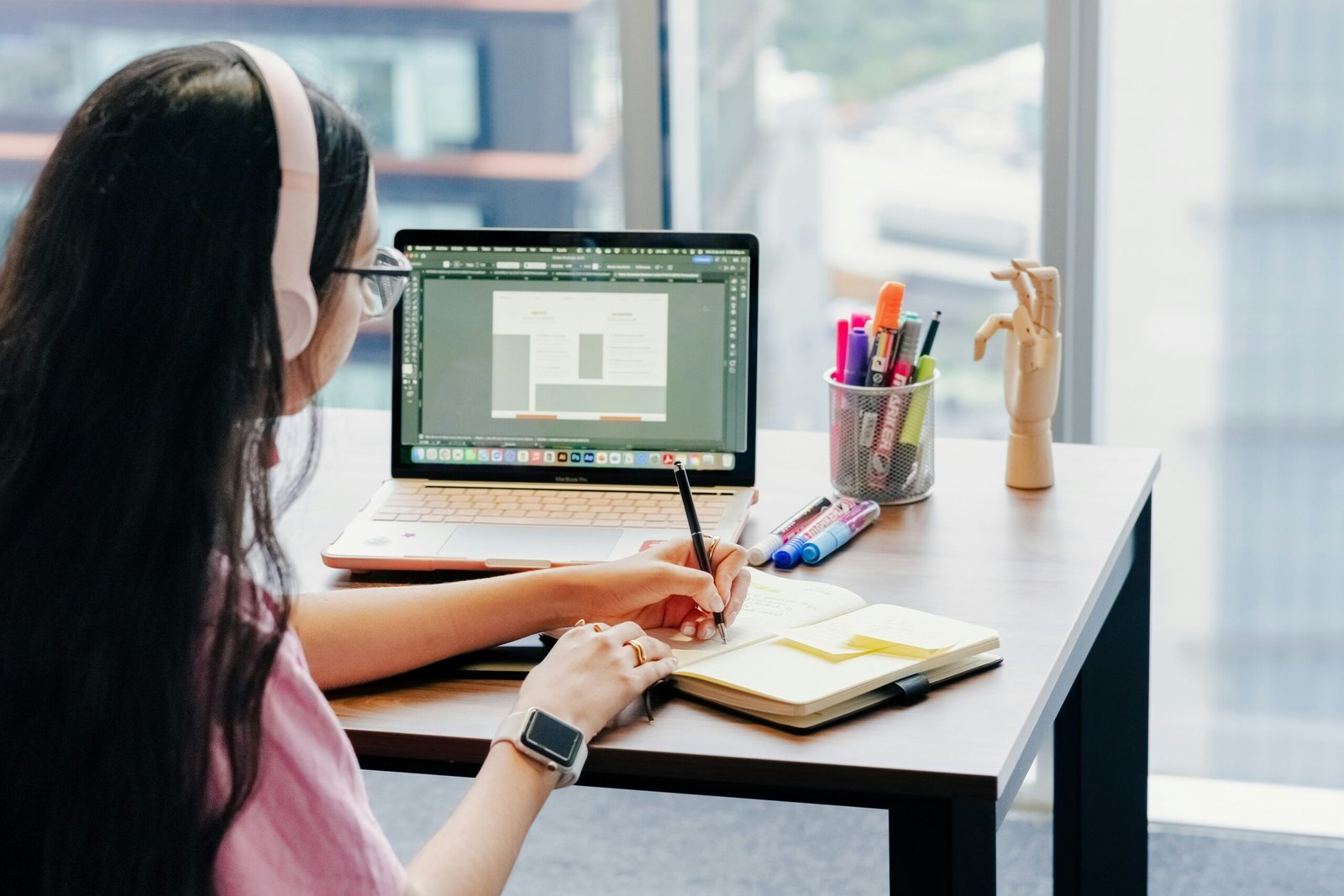A young girl infront of the computer with headphones attending classes and writing notes.