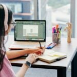 A young girl infront of the computer with headphones attending classes and writing notes.